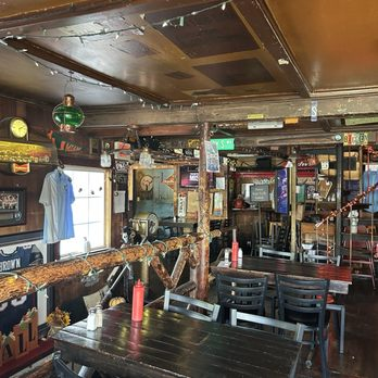 Inside The Dudley Chateau: a long wooden bar rail, dark wood ceiling, framed photographs and neon signs covering the walls, tables with black chairs.