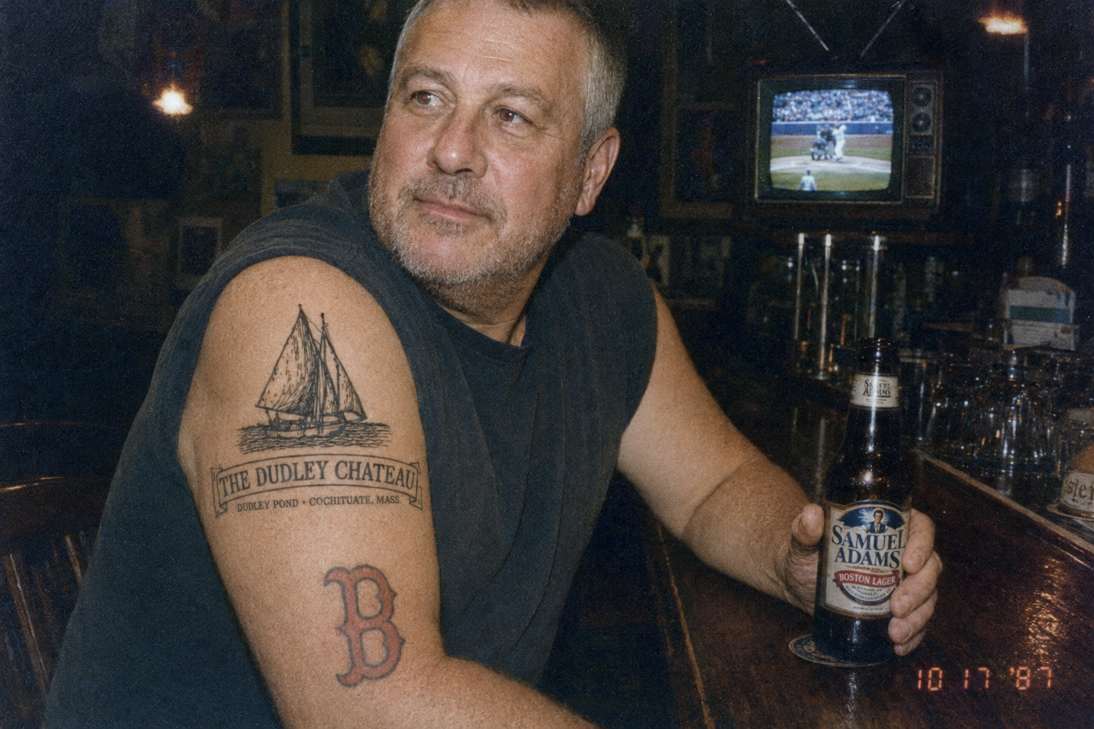 Fictional concept image: a man at a bar with a Sam Adams bottle, the Dudley Chateau schooner logo tattooed on his arm above a Boston B, a ballgame on the television behind him.